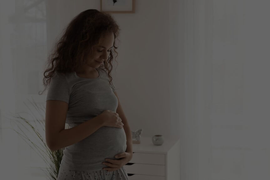 A pregnant woman in a gray shirt stands in a softly lit room, gently holding her belly.